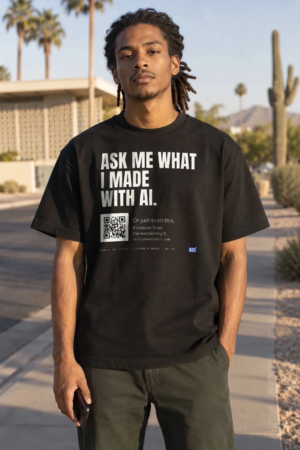 A young man with locs on a sunny Phoenix sidewalk, in a black tee printed: Ask me what I made with AI, with a QR code.