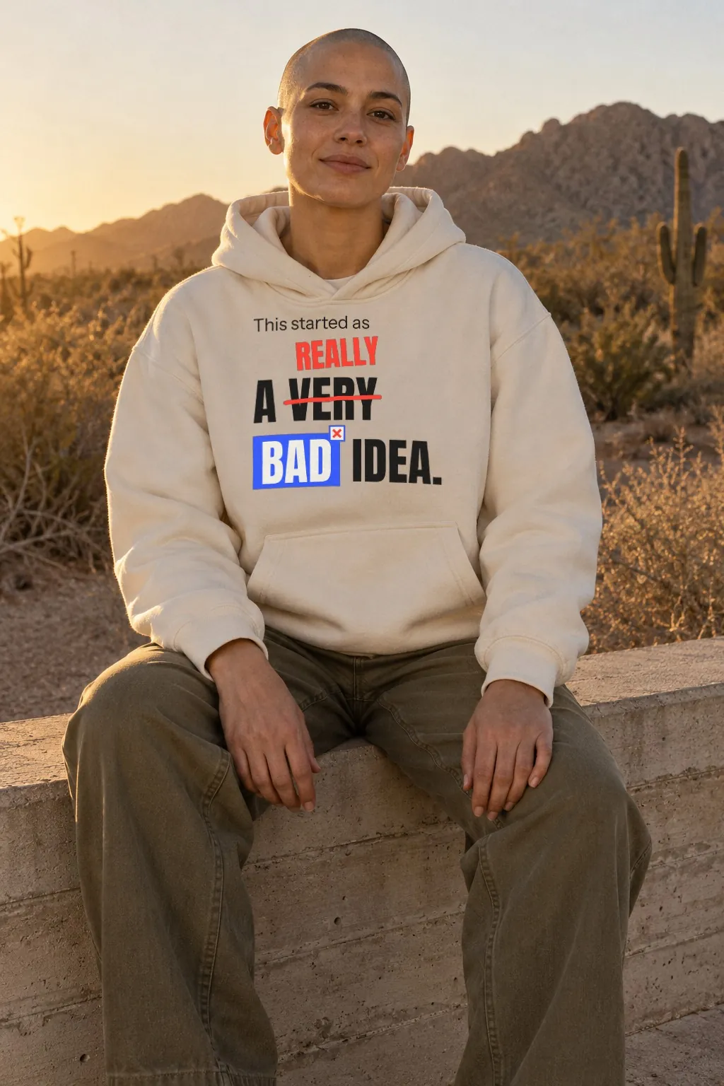A person with a shaved head sitting on a concrete wall at sunset in a bone hoodie printed: This started as a very bad idea, with very crossed out and really written above it.