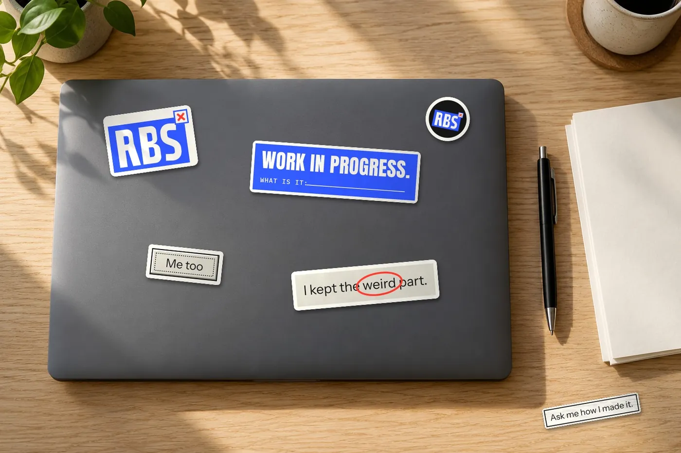 A laptop lid on an oak desk covered in RBS stickers: the RBS tab, Work in progress, Me too, and I kept the weird part.